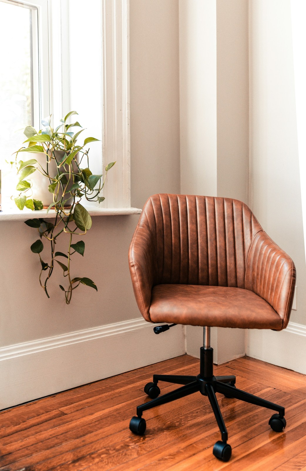 Trailing houseplant on a bright windowsill beside a brown leather desk chair in a quiet room corner